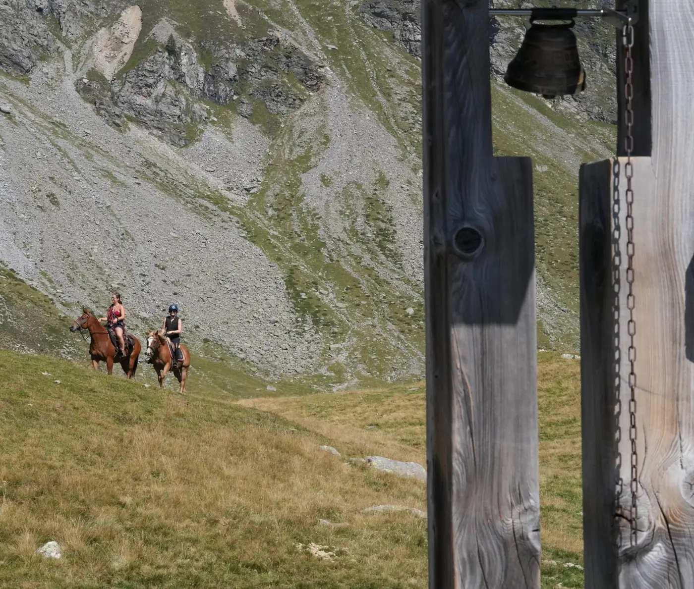 Due persone a cavallo attraversano un prato d'alpe sotto un pendio di roccia, con una campana appesa a un palo di legno in primo piano